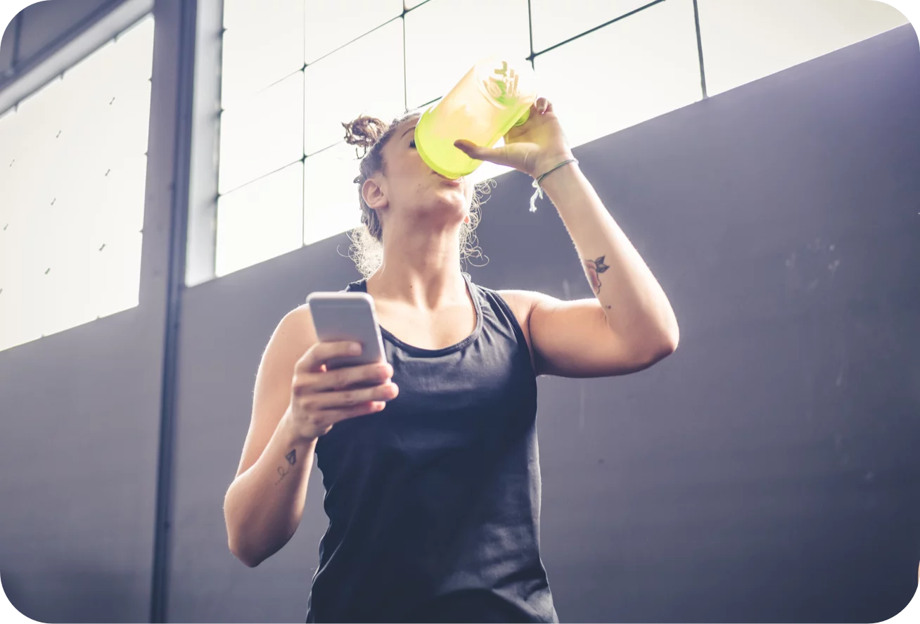 Woman drinking from a yellow water bottle and using a phone outdoors.